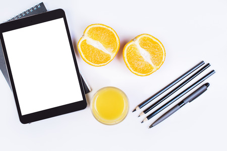 Top view of white desktop with digital tablet, orange juice and fruit halves, pencils and pen. Mock...