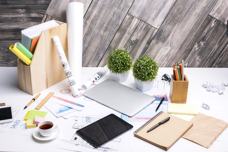 Messy office desktop with empty tablet, coffee cup, documents, decorative plants and other items on...