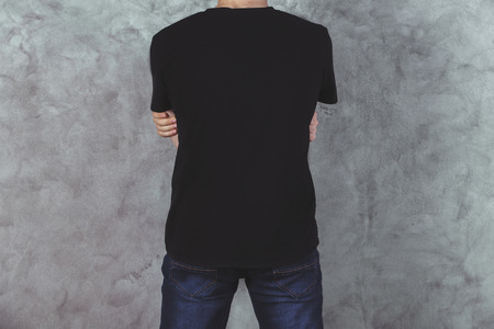 Back view of young man in casual black t-shirt and jeans on concrete wall background. Mock up