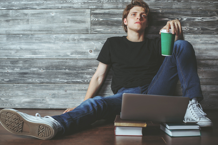 Handsome young boy sitting on wooden floor with books, coffee cup and laptop. Education conceptの写真素材