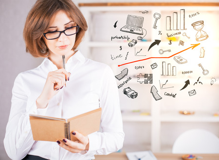 Portrait of attractive caucasian businesswoman writing in notepad with drawn business iconsの写真素材