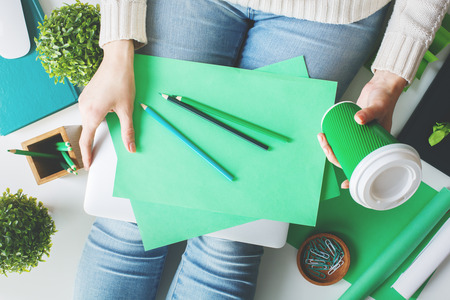 Casual young woman sitting on white desktop with coffee cup, plants, other items and writing on green paper sheetsの写真素材