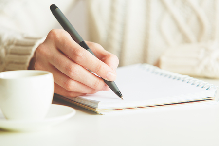 Woman writing in spiral notepad placed on bright desktop with coffee cup. Education concept