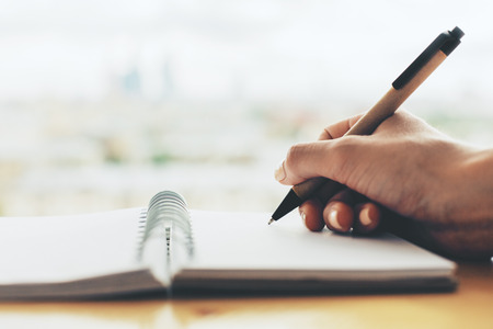 Girl writing in spiral notepad placed on wooden desktop. Blurry background. Close up