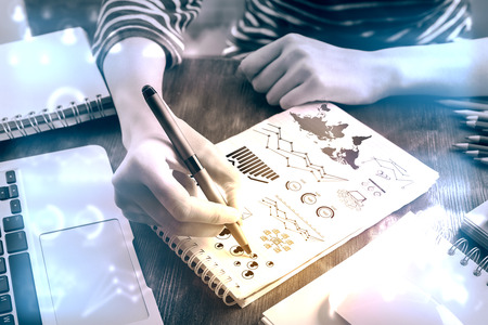 Top view of hands srawing business charts and diagrams on spiral notepad placed on wooden desktpop with laptop and other items. Filtrered imageの写真素材
