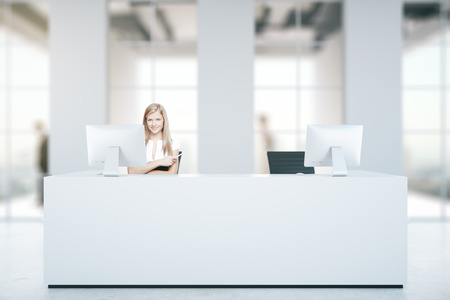 Attractive caucasian woman at white reception desk with computers in blurry concrete interior with daylight. Business concept. 3D Renderingの写真素材