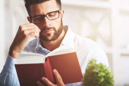 Close up portrait of attractive european man reading book at modern workplace with decorative plant and other items. Education conceptの写真素材