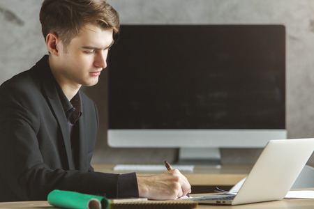 Handsome european man using notebook and doing paperwork at workplace. Blank blurry computer screen in the background. Mock upの写真素材