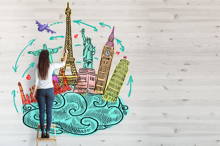 Back view of young woman standing on stool and drawing creative travel sketch on wooden wall. Tourism conceptの写真素材