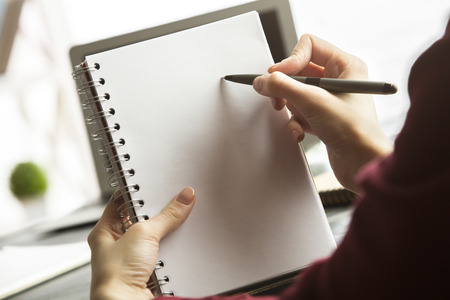 Close up of female hands holding and writing in empty copybook next to office workplace with other supplies and coffee cup. Copy spaceの写真素材