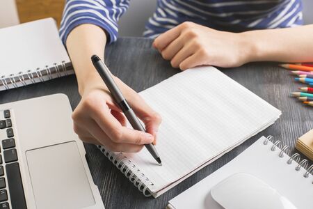Close up of woman's hand writing in empty notepad placed on office desktop with laptop keyboard and...