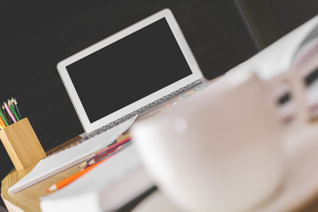 Close up of office desktop with blurry coffee cup and blank laptop screen. Mock up