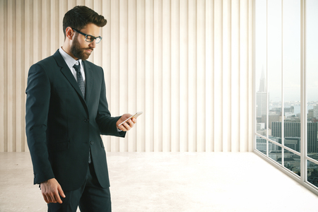 Handsome european businessman using smartphone in interior with city view and copy space on wall. Communication concept. 3D Renderingの写真素材
