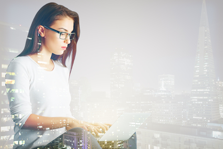Side portrait of attractive businesswoman using laptop on abstract city background with copy space. Communication concept. Double exposureの写真素材