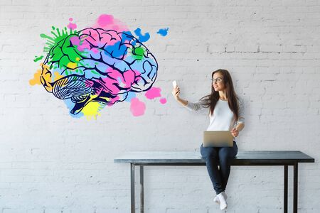 Young woman sitting on table, using laptop and taking selfie with smartphone on white brick background with colorful brain sketch. Brainstorm conceptの写真素材
