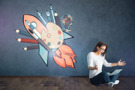 Young european woman sitting on floor and using laptop in dark interior with colorful sketch on wall. Artist conceptの写真素材