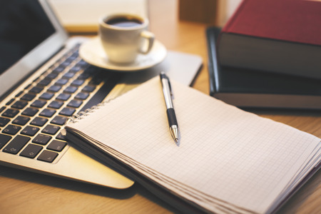 Close up of laptop keyboard, notepad with pen, coffee cup, books and other items on wooden desktop