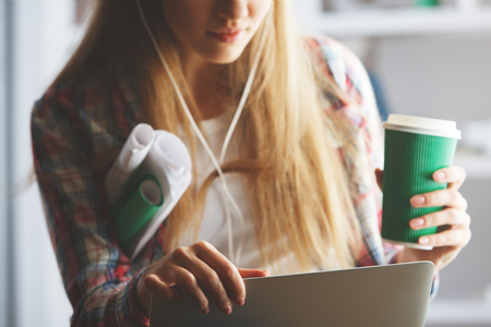 Portrait of attractive young girl with coffee cup and laptop in hands, listening to music through headphonesの写真素材