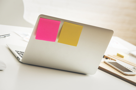 Back view of open laptop with stickers, smartphone, notepad and paperwork placed on white office tableの写真素材