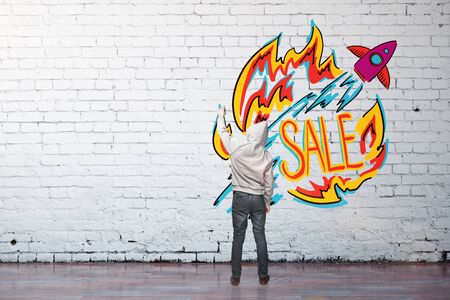 Back view of young guy drawing colorful sketch on white brick wall. Sale conceptの写真素材
