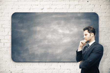 Thoughtful young businessman standing next to empty chalkboard on white brick wall background. Knowledge and science concept. Mock upの写真素材