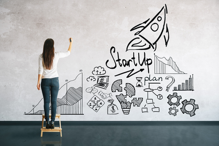 Back view of young woman on stool drawing business sketch on concrete wall. Startup conceptの写真素材