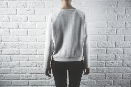 Back view of young woman in casual sweater. Retail concept. White brick wall background