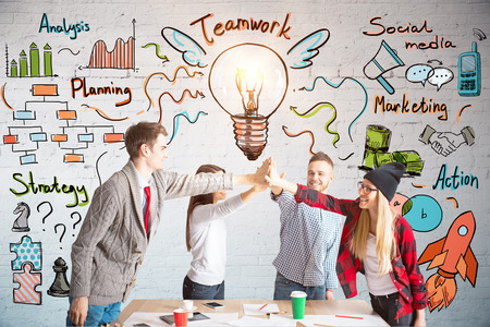Young european team members hi-fiving each other above wooden desk. Brick wall with business sketch...