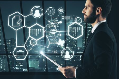 Side view of handsome young businessman with document in hand and abstract digital business icons standing in interior with night city view. Touchscreen concept. 3D Renderingの写真素材