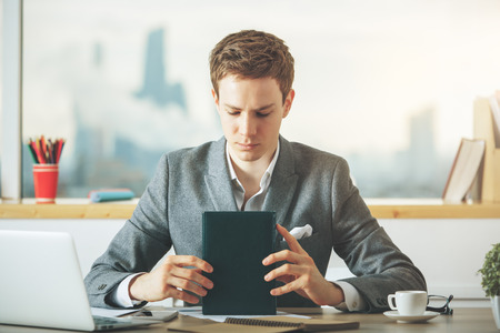 Attractive young european businessman reading book at desktop with laptop, coffee cup and other items in interior with city view. Knowledge conceptの写真素材