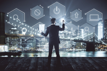 Back view of businessman drawing abstract digital business icons on pier. Illuminated night city background. Infograph screen concept. Double exposureの写真素材