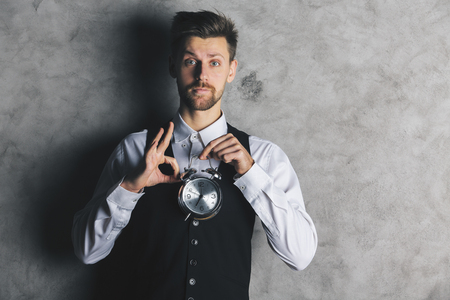 Attractive european businessman holding silver alarm clock and showing ok gesture on concrete backgroundの写真素材