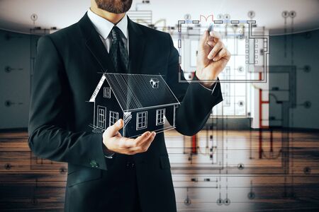Businessman holding abstract house model and drawing on blurry interior background. Architecture concept. 3D Renderingの写真素材
