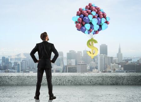 Back view of young businessman on concrete rooftop looking at balloons with golden dollar sign on city background. Profit concept. 3D Renderingの写真素材