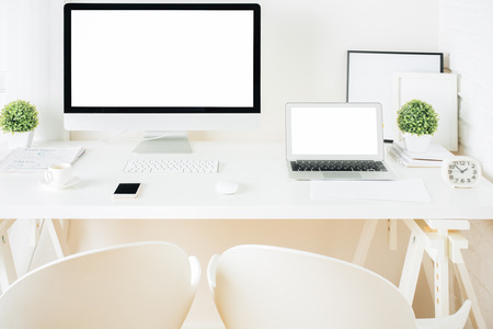 Front view of creative white hipster desktop with empty computer and laptop screens, coffee cup, frames, supplies and other items. Mock upの写真素材