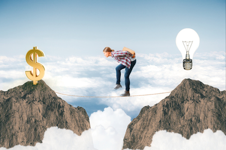 Side view of young woman carrying something on her bag on tight rope between cliffs with dollar sign and lamp. Sky background. Creative business ideasの写真素材