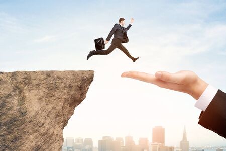 Side view of businessman jumping from cliff to hand on bright city background. Risk and help conceptの写真素材