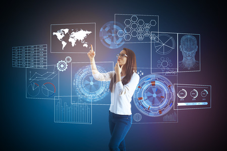 Side view of young woman pointing at abstract futuristic digital business screen. Dark blue background. Future and network concept. Double exposureの写真素材