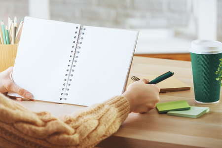 Close up of female hands writing in white spiral notepad at wooden office desk with takeaway coffee cup, decorative plant and other supplies. Mock upの写真素材