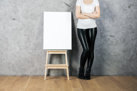 Woman with empty poster on stool standing in room with concrete wall and wooden floor. Advertising concept. Mock upの写真素材