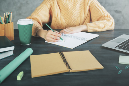 Close up of young woman working on project at wooden hipster office desk with supplies, coffee cup, books, laptop, decorative plant and other items. Workplace and lifestyle conceptの写真素材