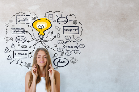 Young woman with crossed fingers on concrete wall background with business sketch. Education and...