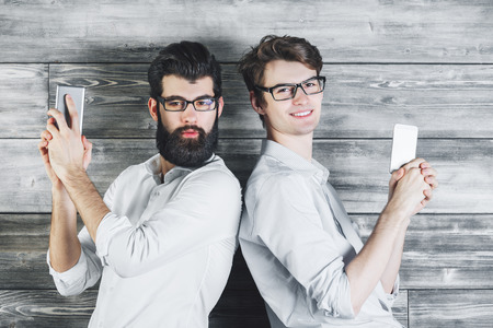 Two handsome businessmen playing with gun smartphones on wooden wall background. Leisure, break, fun conceptの写真素材