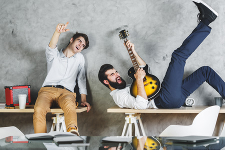European men playing with guitar in office interior with desk and concrete wall background. Leisure, music, rehearsal, band conceptの写真素材