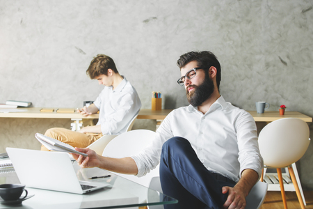 Side view of thoughtful young businessman using laptop in modern office with blurry colleague in the background. Project conceptの写真素材
