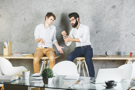 Two handsome businessmen sitting on table and having fun together in modern office. Break conceptの写真素材