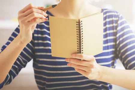 Female in striped blue shirt holding and pointing at empty brown spiral notepad with pen. Mock upの写真素材
