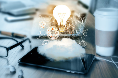 Smartphone with cloud, business hologram and glowing lamp placed on office desktop with coffee cup, supplies and other blurry items. Innovation, cloud computing conceptの写真素材