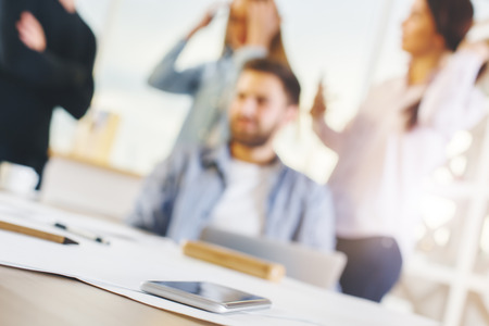 Close up of smartphone on desktop with supplies, coffee cup and other items. Blurry people working in the background. Technology conceptの写真素材