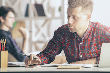 Portrait of handsome caucasian guy writing in spiral notepad at workplace. Paperwork concept. Blurry colleague in the backgroundの写真素材